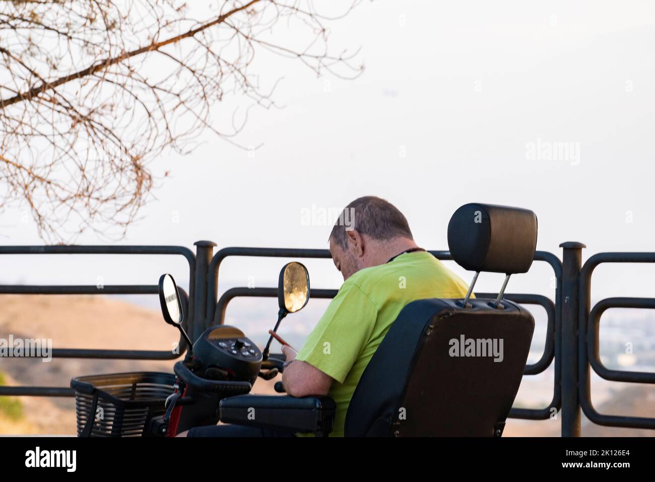 Ein behinderter Erwachsener sitzt in einem motorisierten Rollstuhl an einem Aussichtspunkt und blickt auf sein Mobiltelefon. Stockfoto