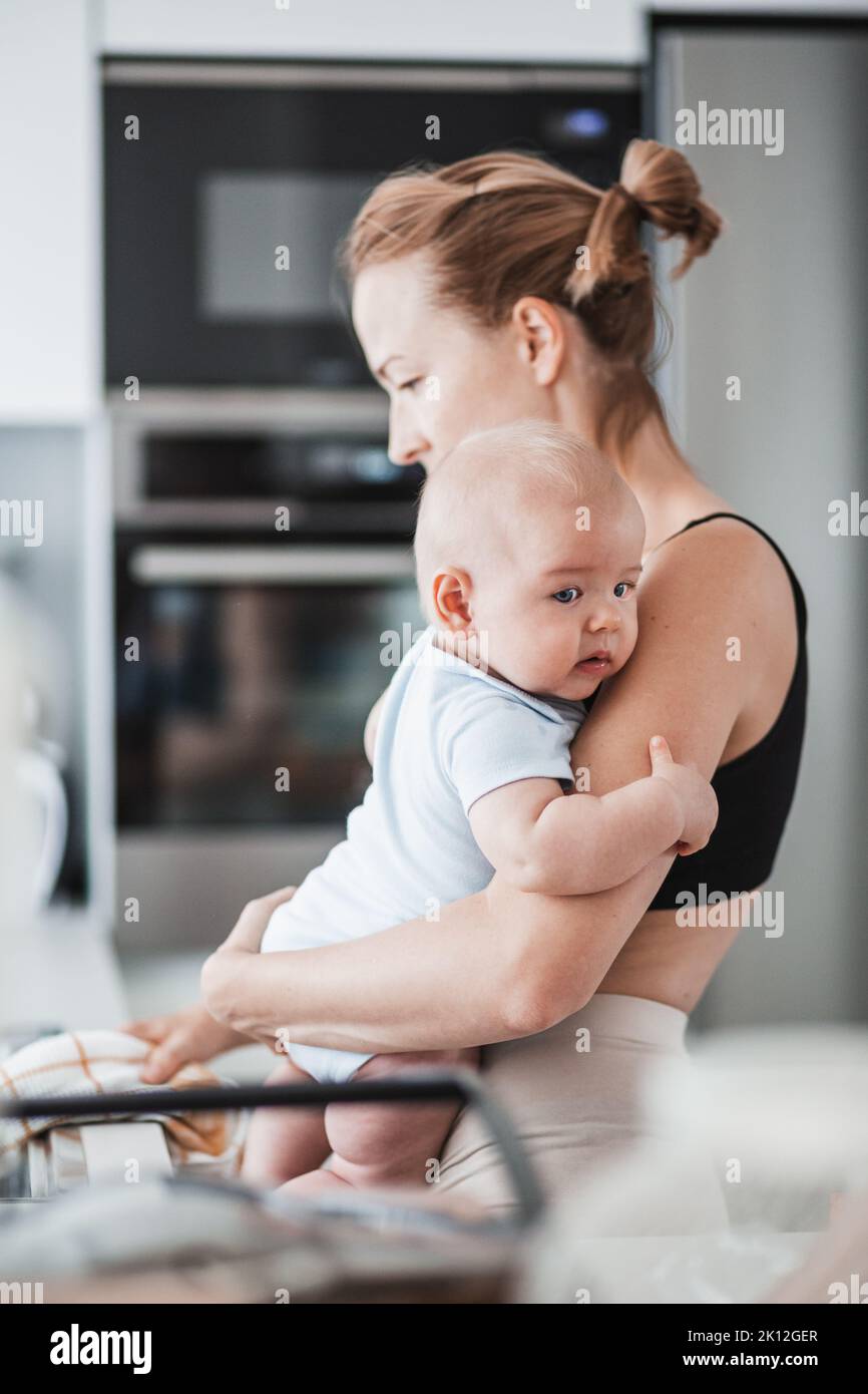 Frau wischt nach dem Abwaschen das Spülbecken mit einem Tuch ab, während sie den vier Monate alten Jungen in den Händen hält Stockfoto