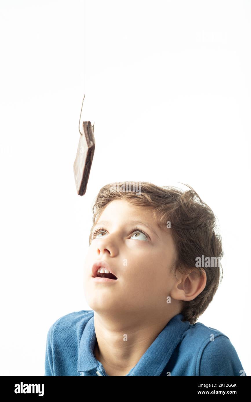 Ein Kind blickt sehnsüchtig auf einen Biskuitkuchen, der an einem Haken hängt. Das Bild zeigt das Konzept der Suchtproblematik gegen Zucker und süße Lebensmittel. Weißer Hintergrund. Stockfoto