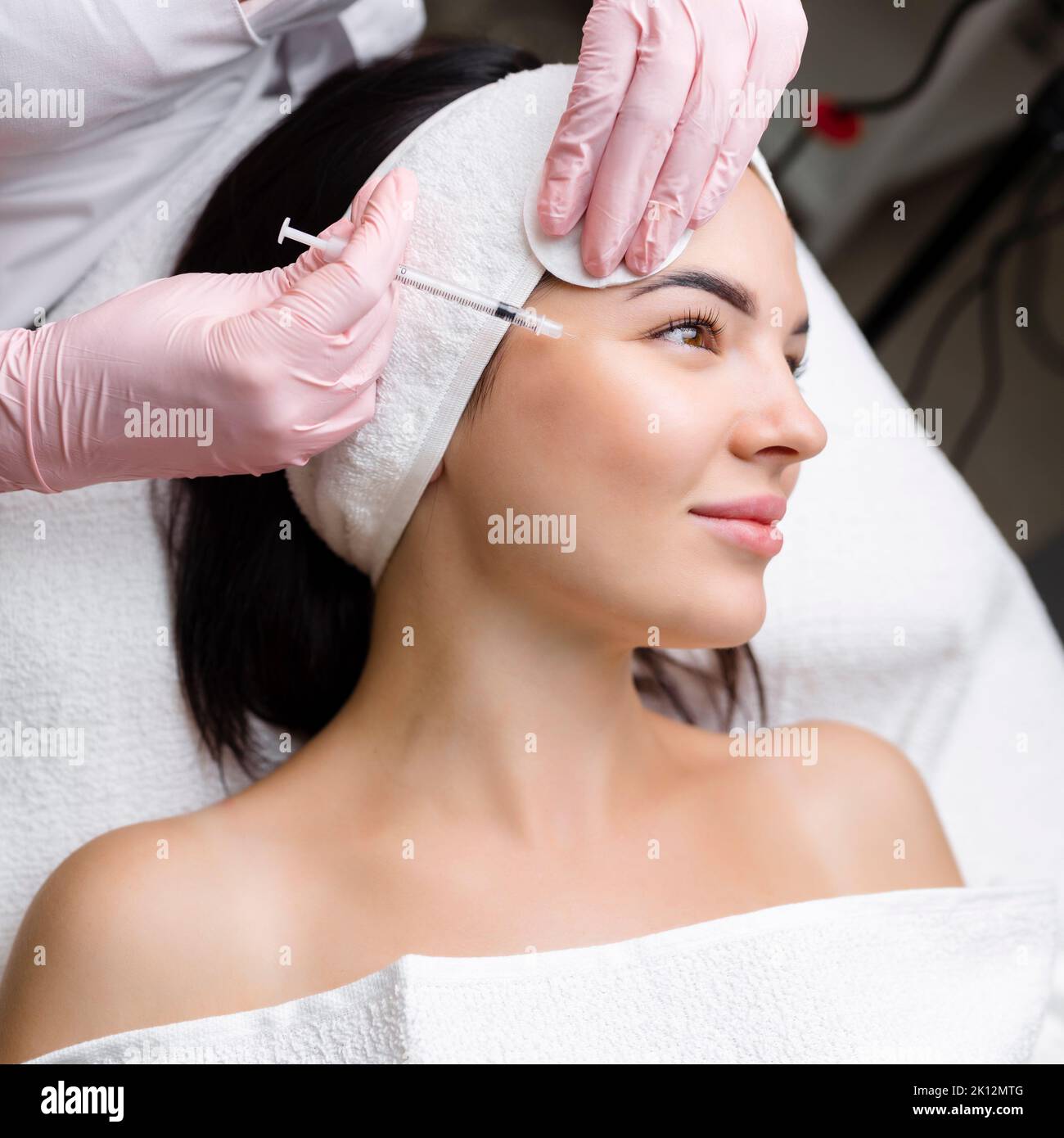 Nahaufnahme der Hände eines jungen Kosmetologen, der Botox in das weibliche Gesicht injiziert. Sie steht und lächelt. Die Frau schließt ihre Augen mit Entspannung Stockfoto