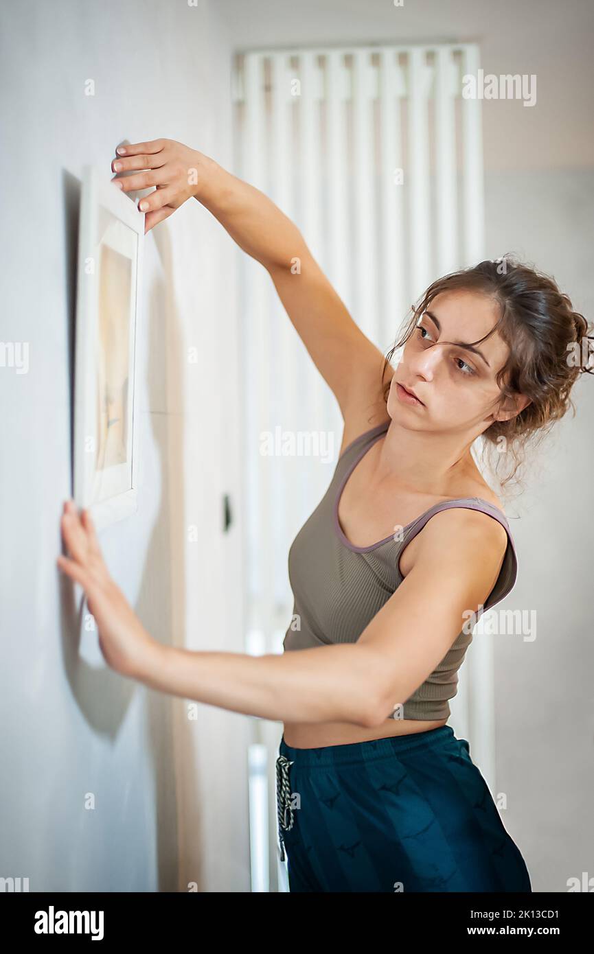 Fröhliche junge Frau schmückt ihre neue Wohnung mit Bildern an der Wand Stockfoto