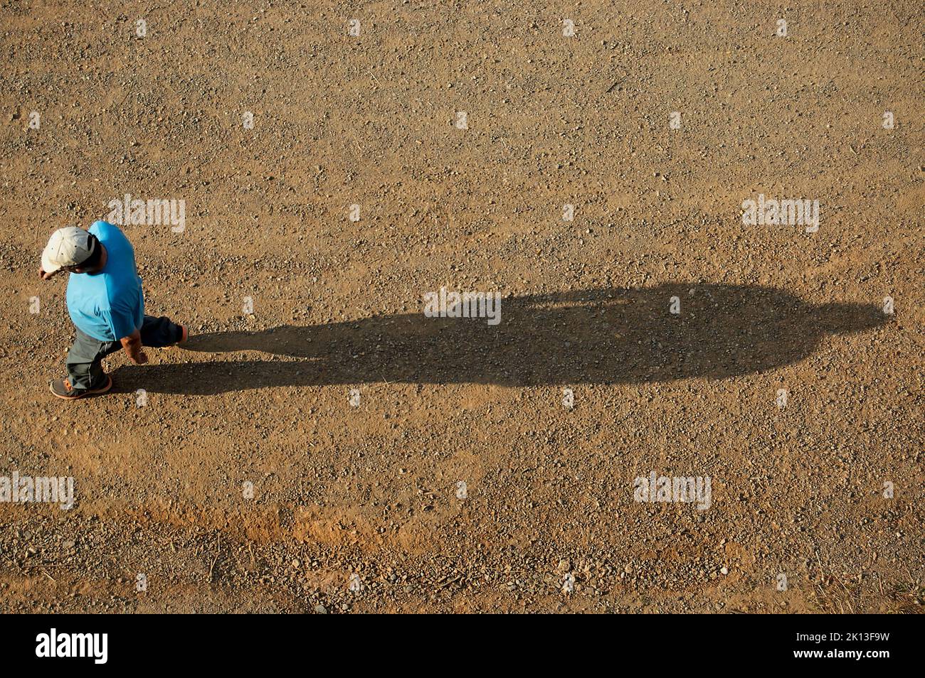 Landmann in kurzen Ärmeln mit Mütze und Turnschuhen mit der Sonne vor ihm, der mit seinem Schatten hinter ihm auf braunem schmutzigen Boden und ganz nah spazierend Stockfoto