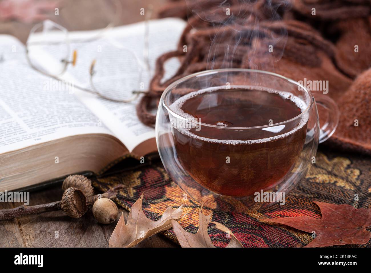 Nahaufnahme von dampfendem heißen Tee in einer Glastasse mit Buch, Brille und Decke im Hintergrund Stockfoto