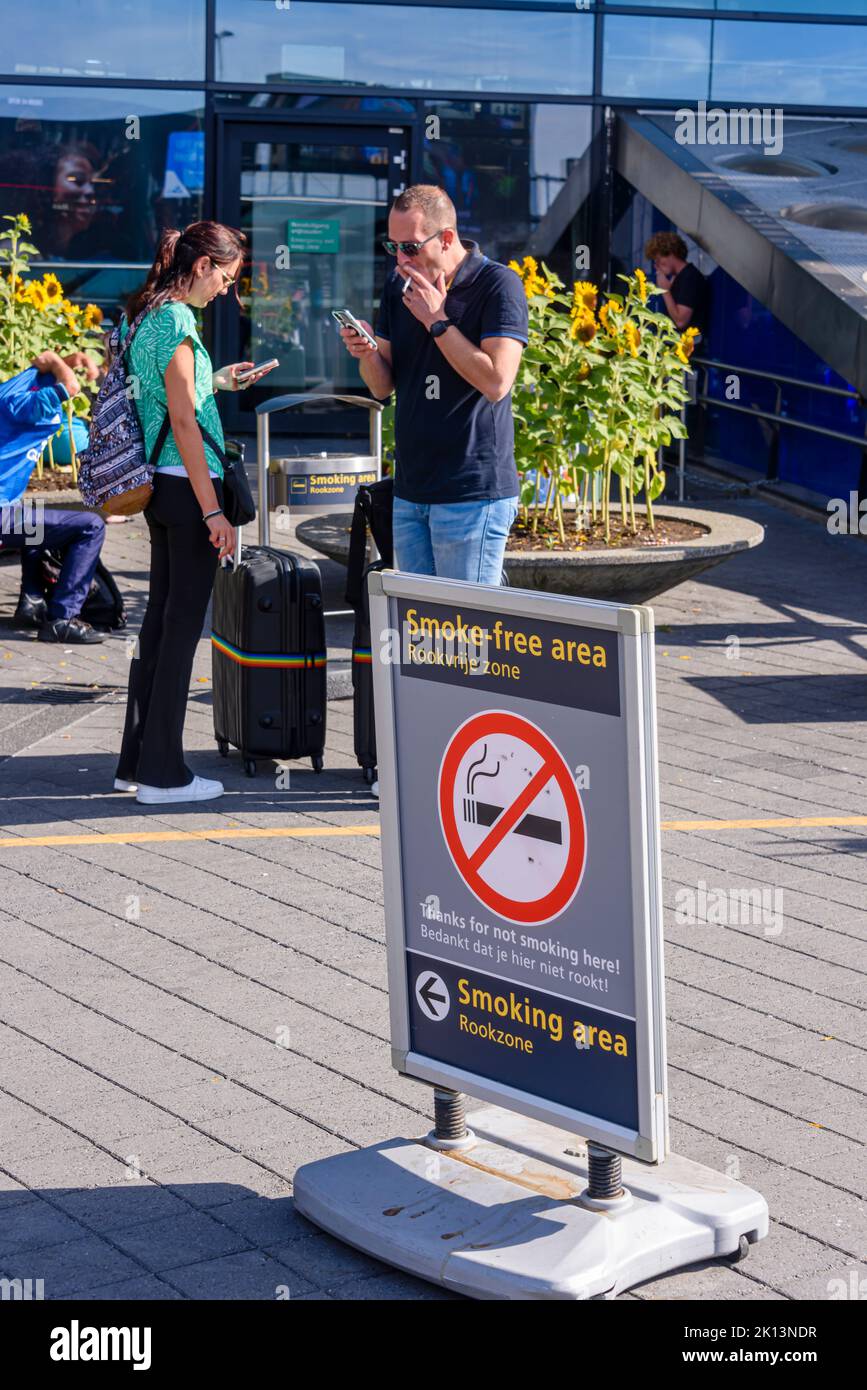 Zwei Personen rauchen hinter einem Schild, auf dem steht, dass es sich um einen rauchfreien Bereich handelt, und zeigen auf einen Raucherbereich, den Flughafen Schiphol, Niederlande Stockfoto