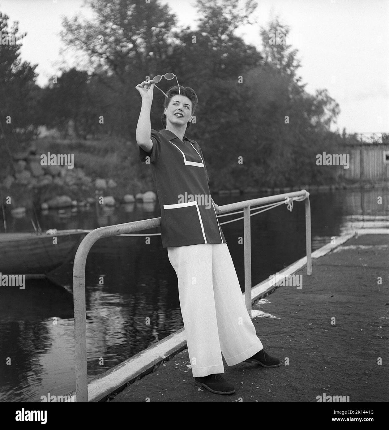 Damenmode im Jahr 1940s. Eine junge Frau, die für ein Damenmagazin fotografiert wurde, um die Damenmode von 1945 zu zeigen. Das weibliche Modell trägt eine weite Hose und eine kurze Bluse oder Jacke. Sie sieht ihre Sonnenbrille gegen den Himmel an, als ob sie sie reinigen muss. Schweden 1945. Kristoffersson Ref. R6-2 Stockfoto