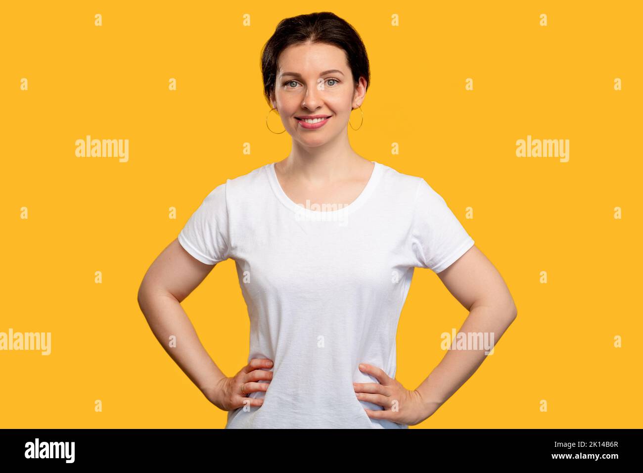 Selbstbewusstes Frauenporträt. Lässiger Style. Fröhliche Dame im weißen T-Shirt mit Händen an der Hüfte. Blick auf die Kamera isoliert auf orange. Erfolgreicher Erfolg. W Stockfoto