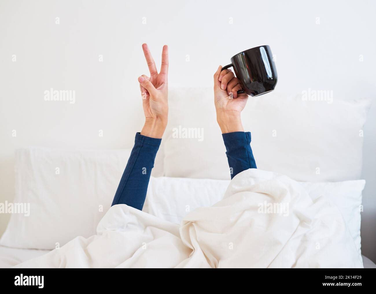 Nahaufnahme der Hände, die ein Friedenszeichen bilden, mit einem Kaffeetasse, der aus der Bettdecke seinen Höhepunkt erreicht Stockfoto