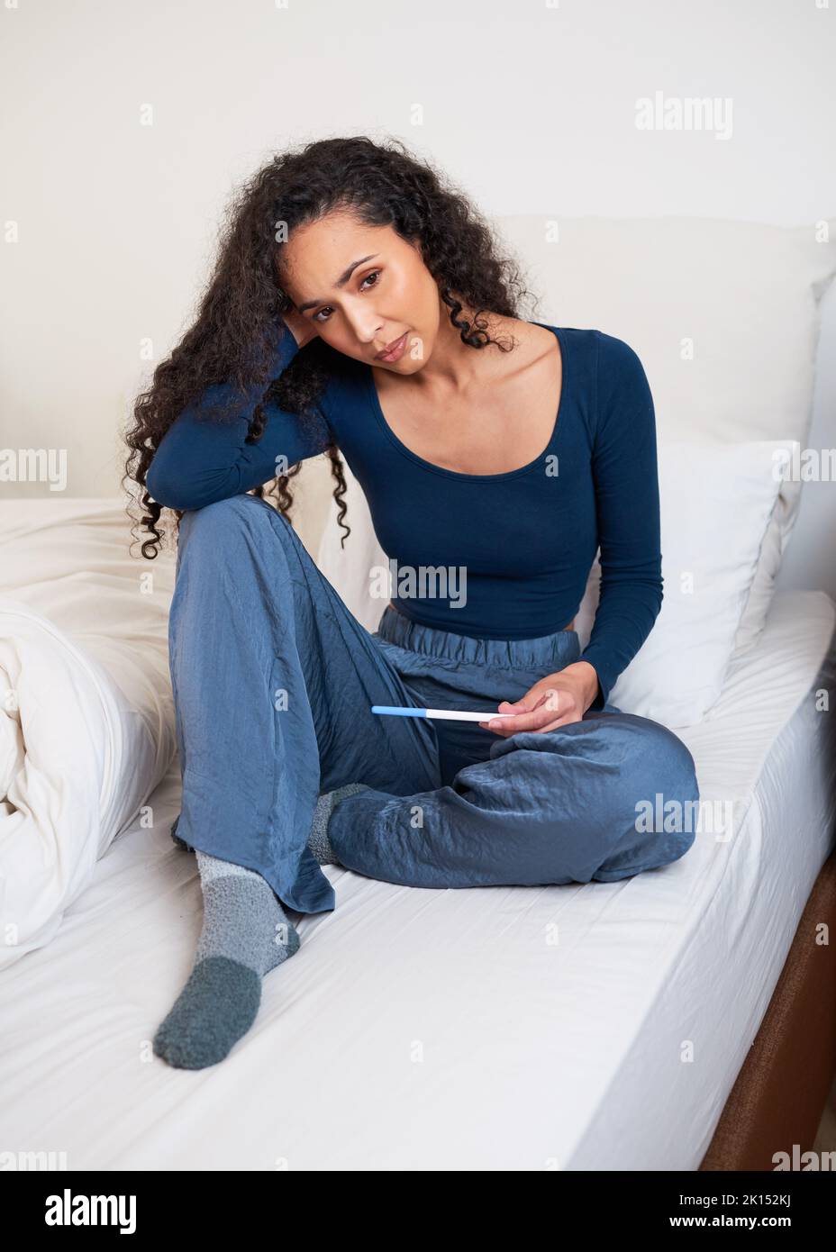 Eine junge, multiethnische Frau sitzt im Bett, verärgert über das unerwünschte Ergebnis eines Schwangerschaftstests Stockfoto