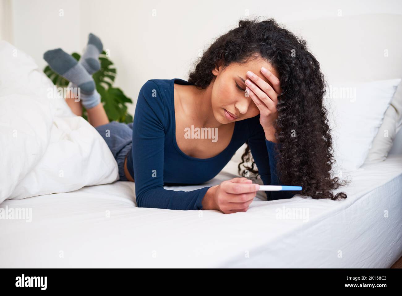 Eine junge, multiethnische Frau liegt im Bett und schaut sich das Ergebnis eines unerwünschten Schwangerschaftstests an Stockfoto