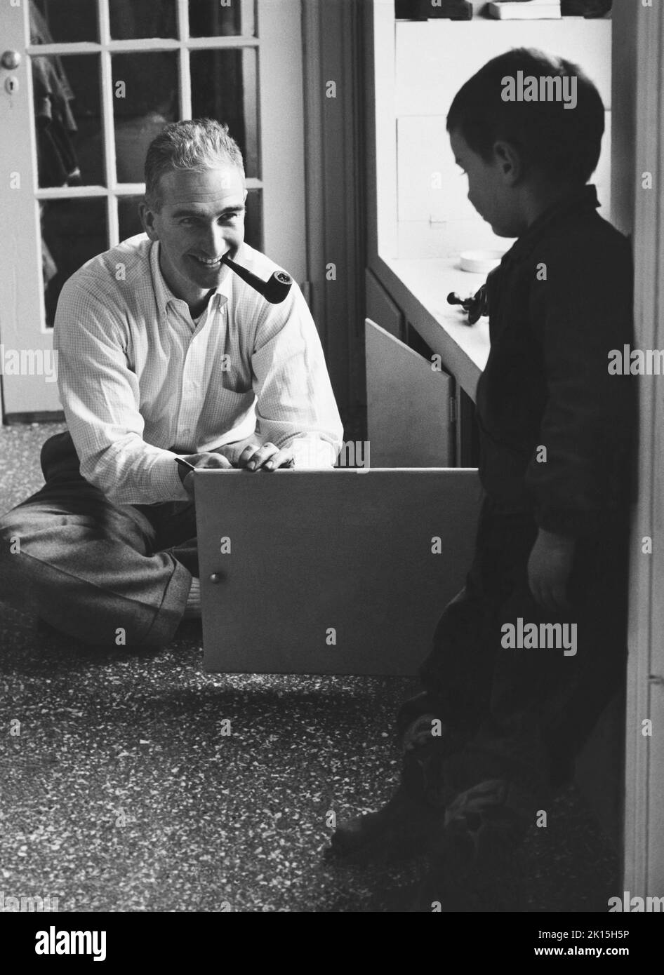 Ein Pfeifenraucher Vater öffnet eine Schranktür, während sein Sohn zuschaut. Ca. 1960. Stockfoto