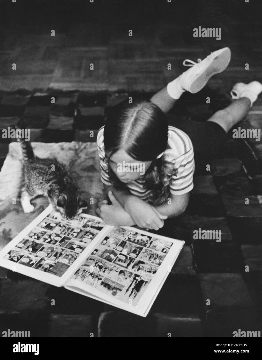 Ein Mädchen und ihr Kätzchen lagen zusammen auf einem Teppich und lasen ein Buch mit Cartoons. Ca. 1960, 1970. Stockfoto