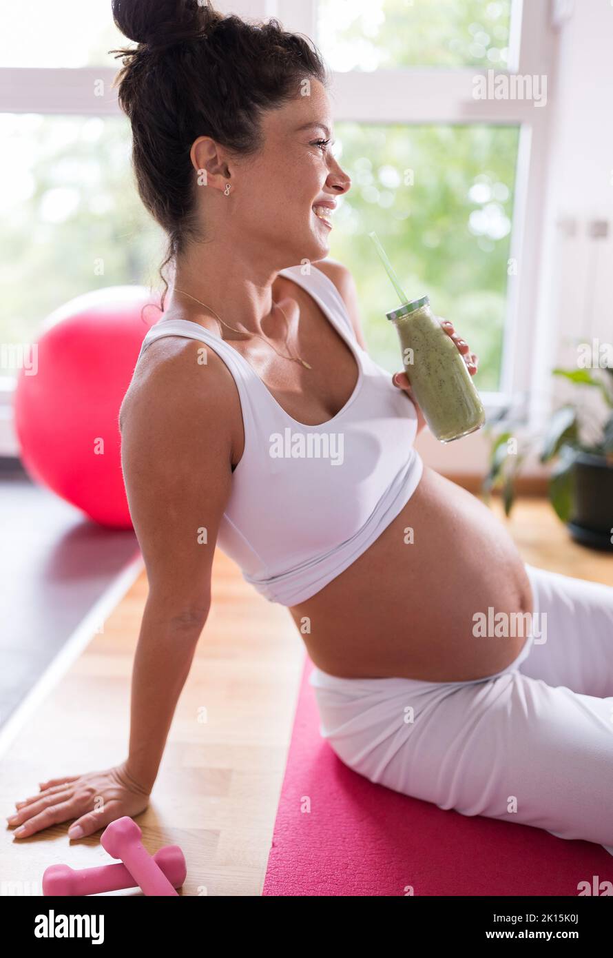 Hübsche Schwangere, die sich nach dem Training auf einer Yogamatte ausruht und grünen Smoothie trinkt. Gesundes Lebenskonzept für die Mutterschaft Stockfoto