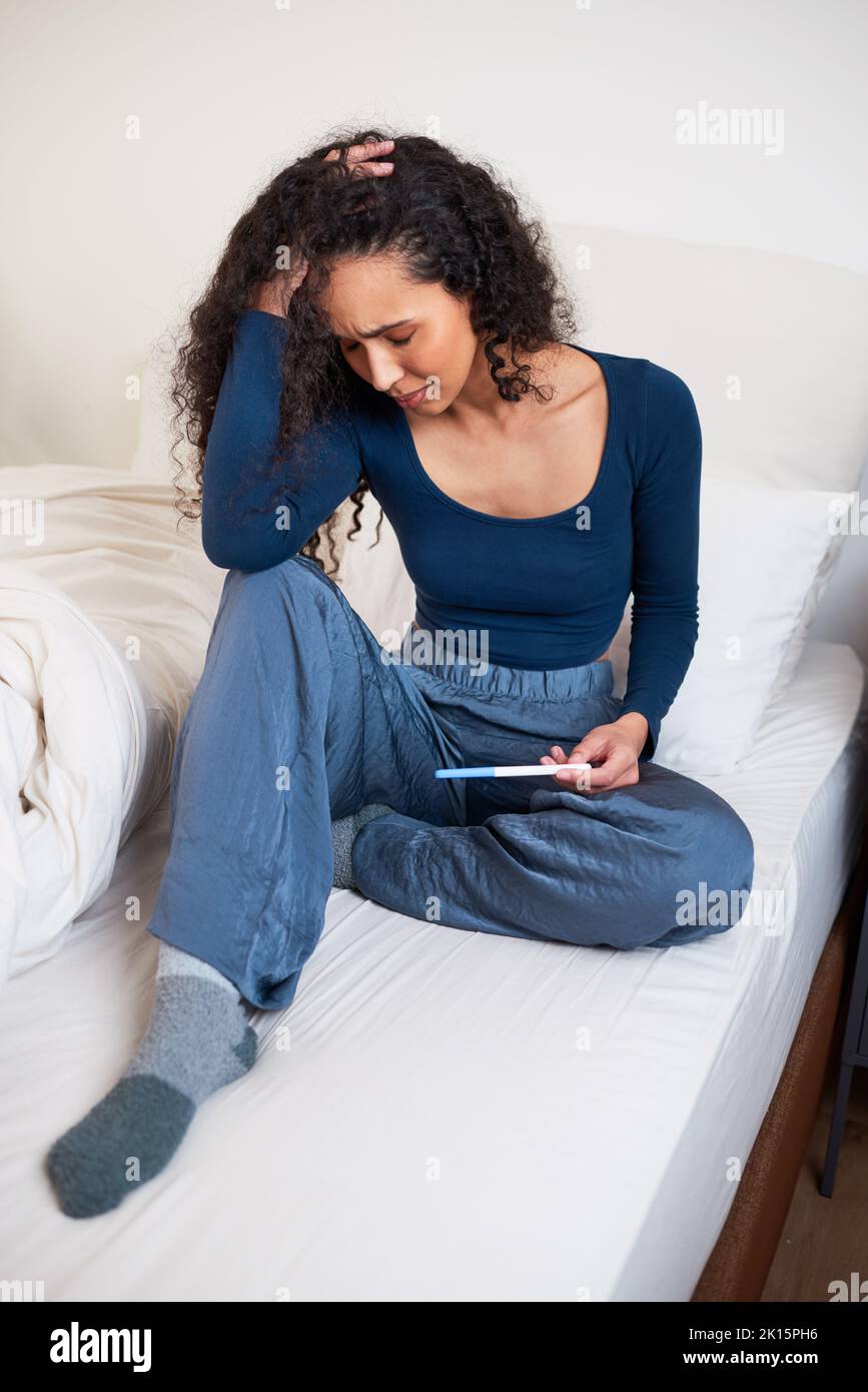 Eine junge, multiethnische Frau sitzt im Bett, verärgert über das unerwünschte Ergebnis eines Schwangerschaftstests Stockfoto