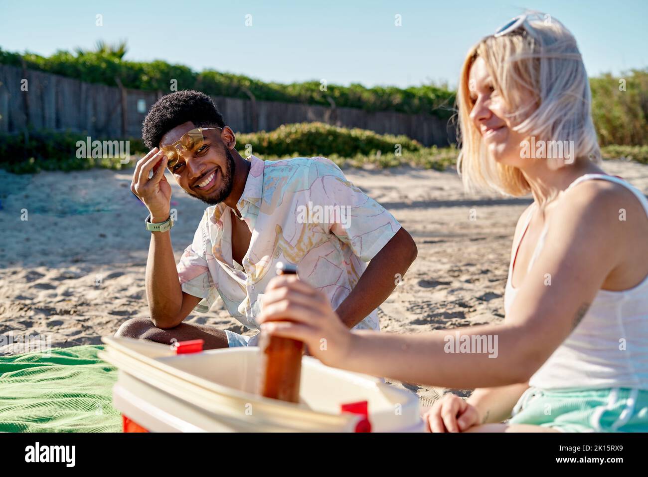 Glücklicher afroamerikanischer Mann, der mit einem Lächeln auf die Kamera blickt und die Sonnenbrille am Kopf anpasst, während er während eines Picknicks neben einer Frau sitzt, die Bier vom Kühler nimmt Stockfoto