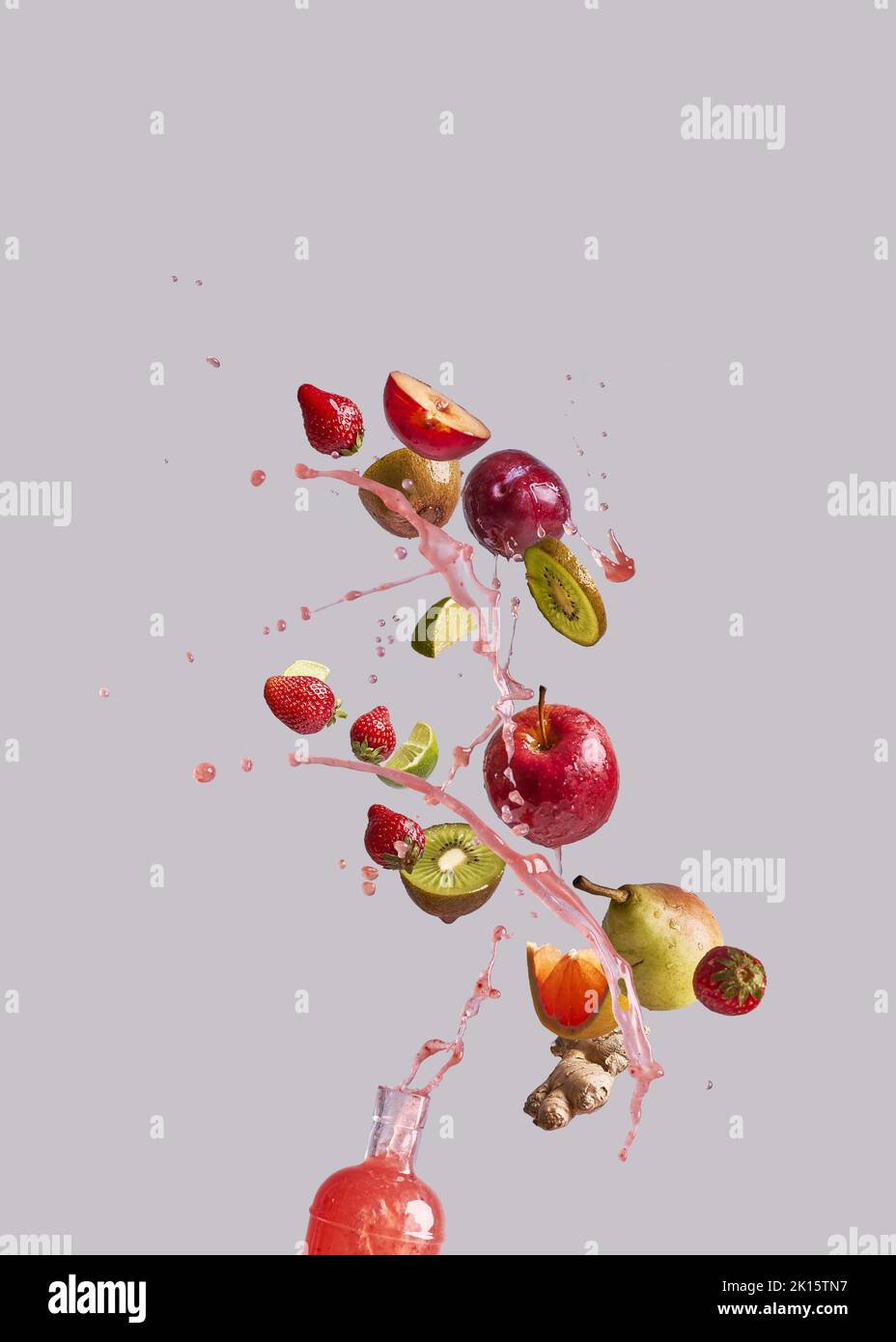 Ein paar verschiedene frische Früchte und Ingwer fallen in eine Glasflasche mit gesundem Saft vor grauem Hintergrund Stockfoto