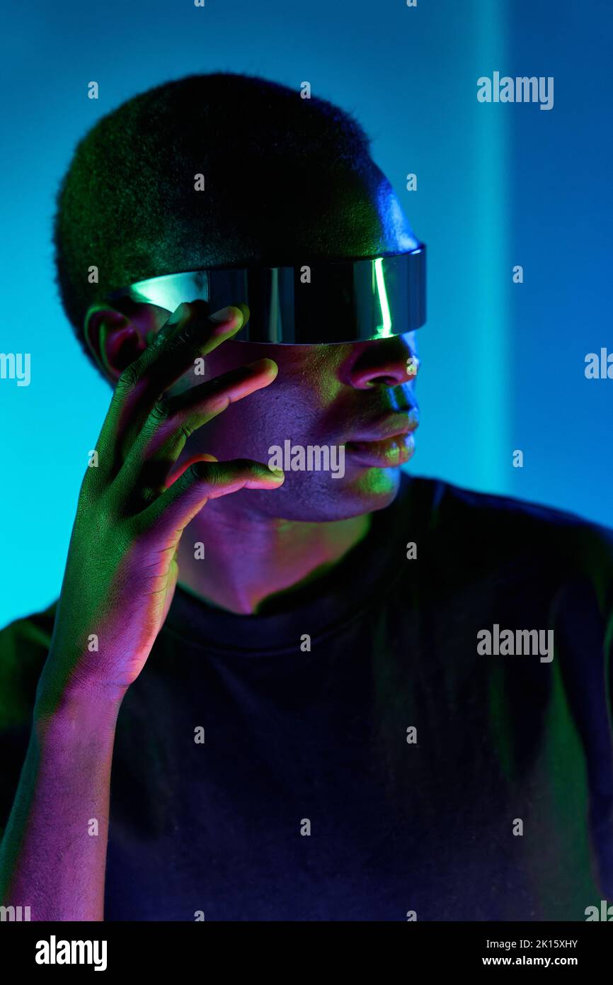 Fokussierter cooler junger Afrikaner in schwarzem T-Shirt, der moderne futuristische Brille anpasst, während er im Studio mit farbenfrohem Ne die virtuelle Realität erlebt Stockfoto