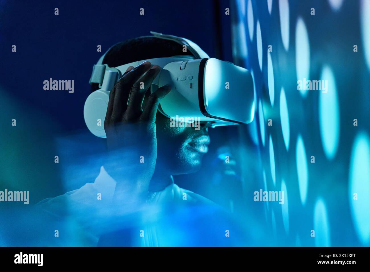 Seitenansicht des konzentrierten jungen Afroamerikaners in weißem T-Shirt und moderner VR-Brille, die mit einem virtuellen Objekt in einem dunklen Studio mit Neon-Illum interagiert Stockfoto