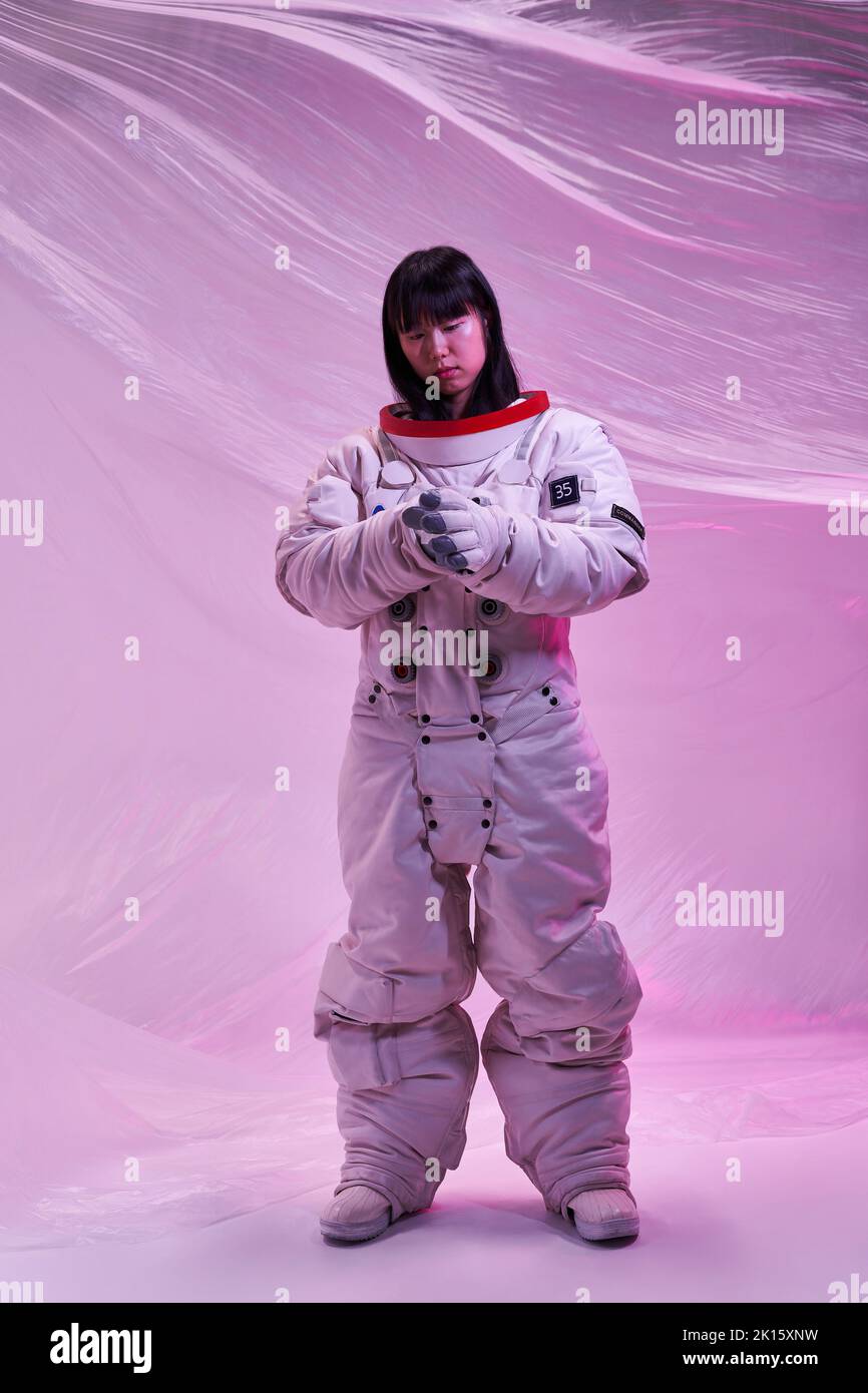 Ganzkörperfrau aus Asien in einem Raumanzügen, die Handschuhe anpasst, während sie unter pinkem Neonlicht vor futuristischem Hintergrund steht Stockfoto