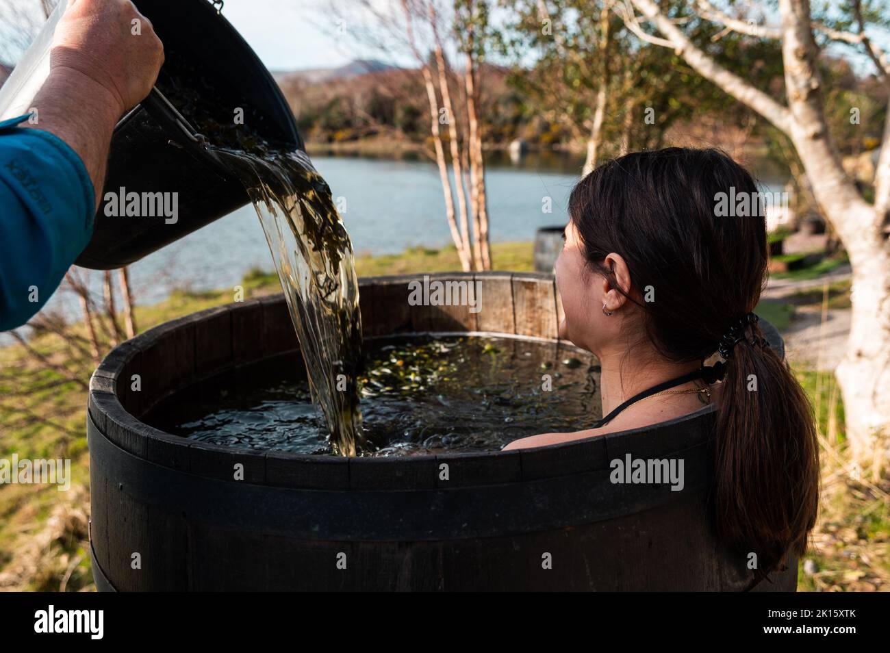 Crop männlichen Gießen Wasser mit Algen in Holzfass mit Brünette heißen Bad am Ufer des Sees im Herbst Stockfoto