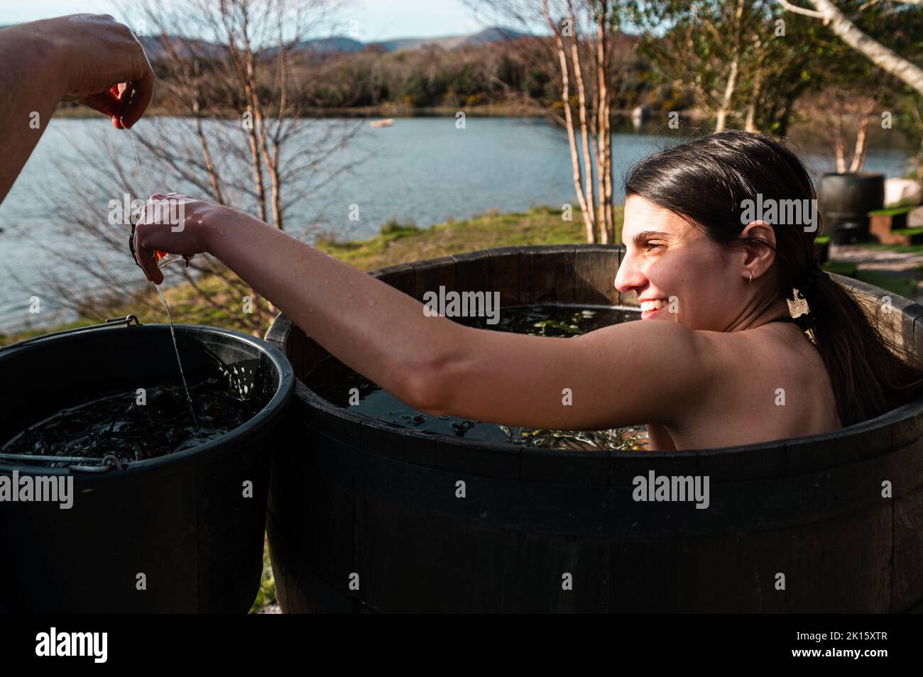 Ernte männlich Wasser mit Algen in Holzfass mit lächelnden Brünette unter heißen Bad am Ufer des Sees im Herbst Stockfoto