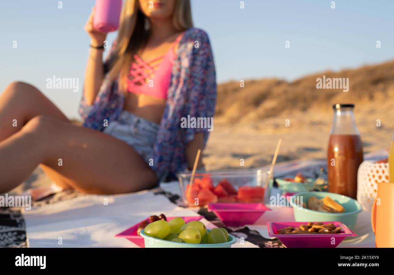 Anonyme, beschnittene Frau mit langen Haaren sitzt auf der Decke und schlürft während des Picknicks am Sandstrand ein frisches Getränk aus dem Becher Stockfoto