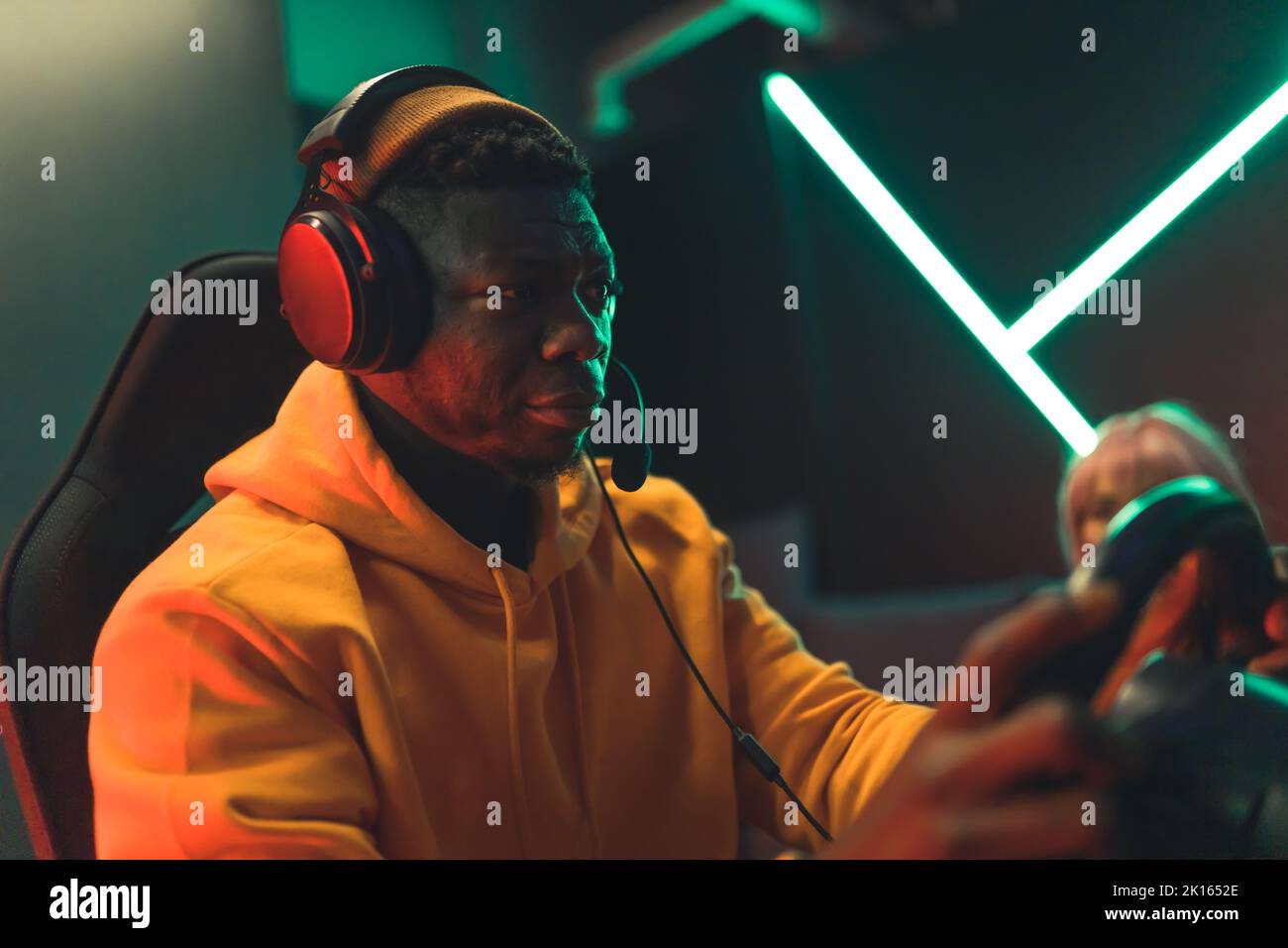 Teenager schwarzer Kerl trägt Headset und orangenen Hoodie spielen Rennspiel mit Lenkrad control. Fokussierter Look. Dunkelgrüne Beleuchtung. Hochwertige Fotos Stockfoto