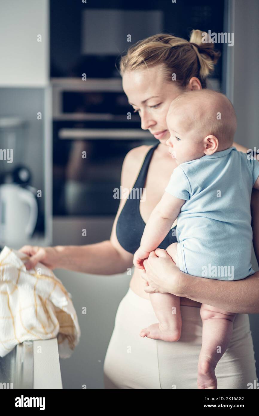 Frau wischt nach dem Abwaschen das Spülbecken mit einem Tuch ab, während sie den vier Monate alten Jungen in den Händen hält Stockfoto