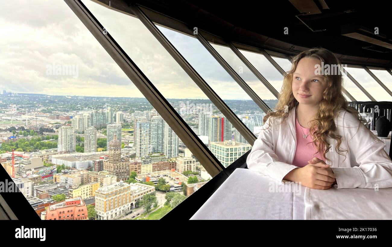Ein junges Mädchen im Teenageralter sitzt am Fenster eines sich drehenden Restaurants und ist traurig, dass sie ihre Hand neben ihrem Gesicht traurig ist und sie aus dem Fenster auf das Visights Top of Vancouver Revolving Restaurant blickt. 4K Stockfoto