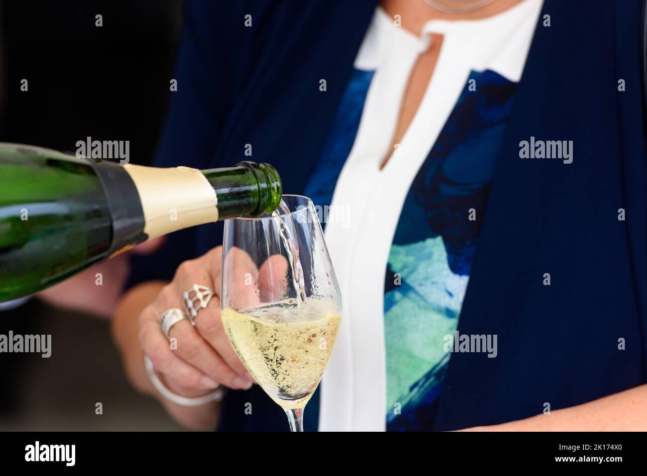Nahaufnahme einer Champagnerflasche, die Champagner auf einer Sektflöte gegen eine verschwommene Person auf einer Party umschaufelt Stockfoto