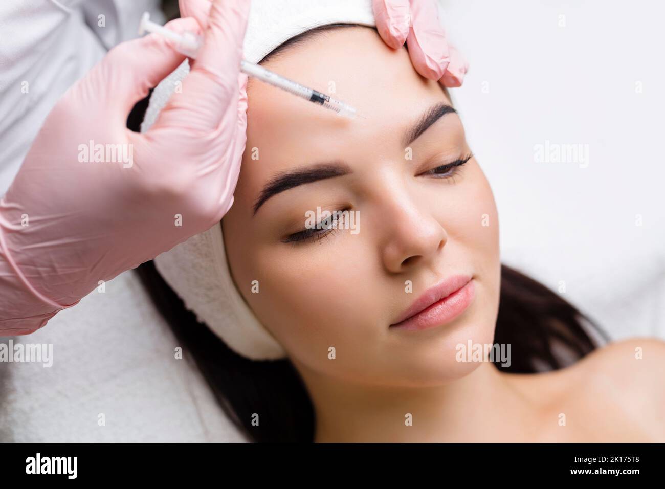 Nahaufnahme der Hände eines jungen Kosmetologen, der Botox in das weibliche Gesicht injiziert. Sie steht und lächelt. Die Frau schließt ihre Augen mit Entspannung Stockfoto