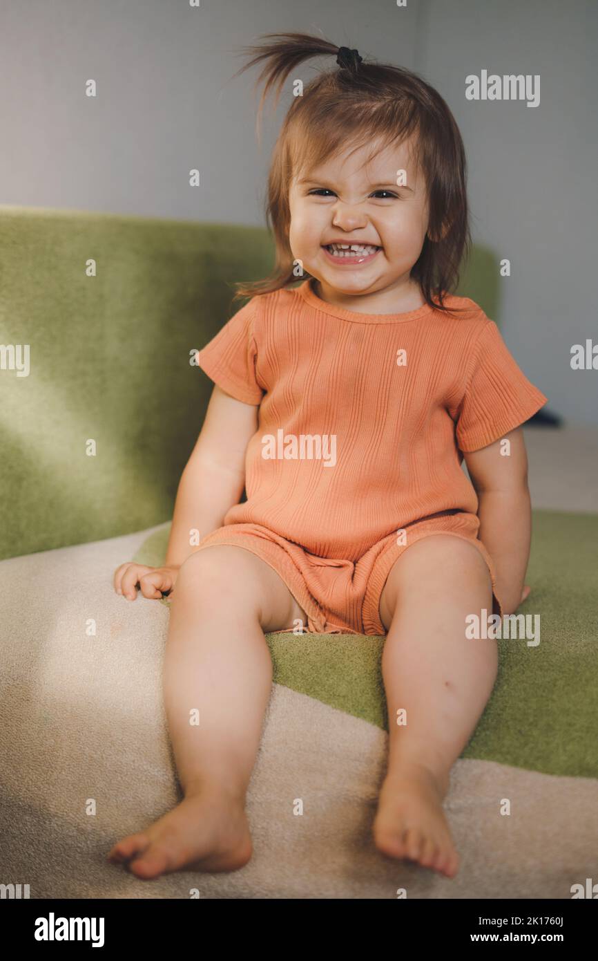 Entzückendes kleines Baby mit orangefarbenem Kostüm, das auf dem Sofa sitzt, lächelnd auf die Kamera schaut. Glückliche Familie, Kindheit. Lächelndes Mädchen. Kinderbetreuung. Stockfoto