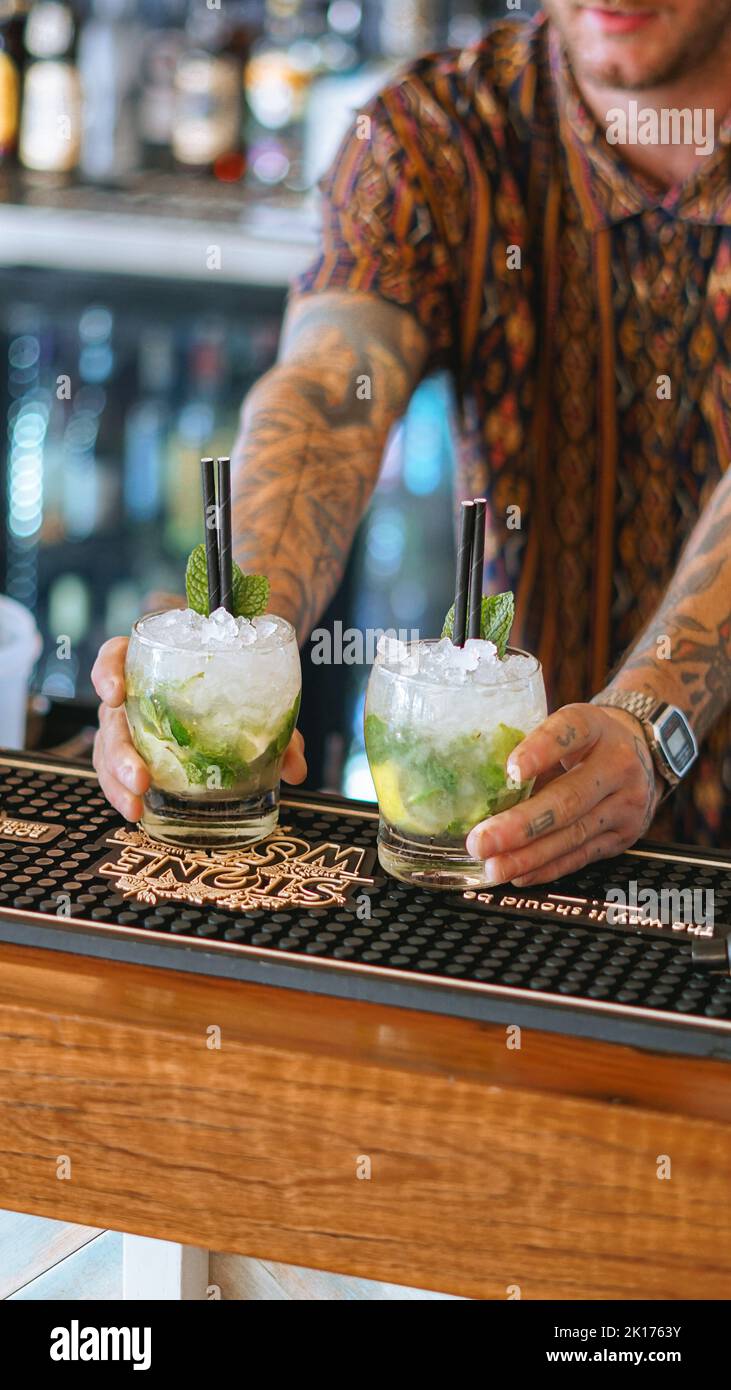 Der männliche Barkeeper serviert 2 Mojito-Cocktails hinter der Bar. Er trägt ein schwarzes Hemd, eine Uhr und serviert die Getränke mit zwei schwarzen Trinkhalmen. Stockfoto