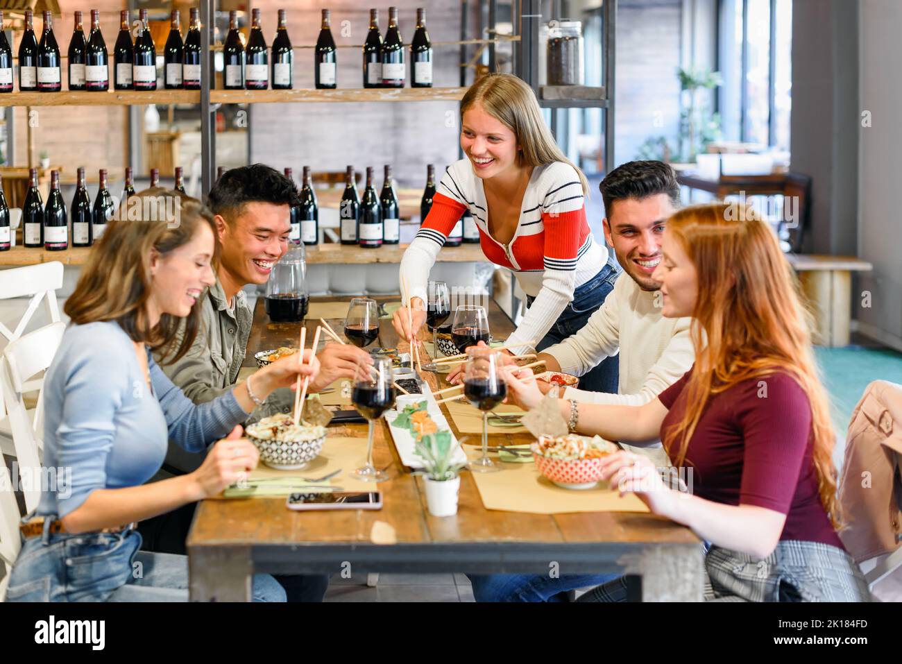 Positive, vielfältige Freunde, die beim gemeinsamen Abendessen am Tisch mit Gläsern Rotwein in einem modernen, hellen Restaurant leckeres Essen essen Stockfoto
