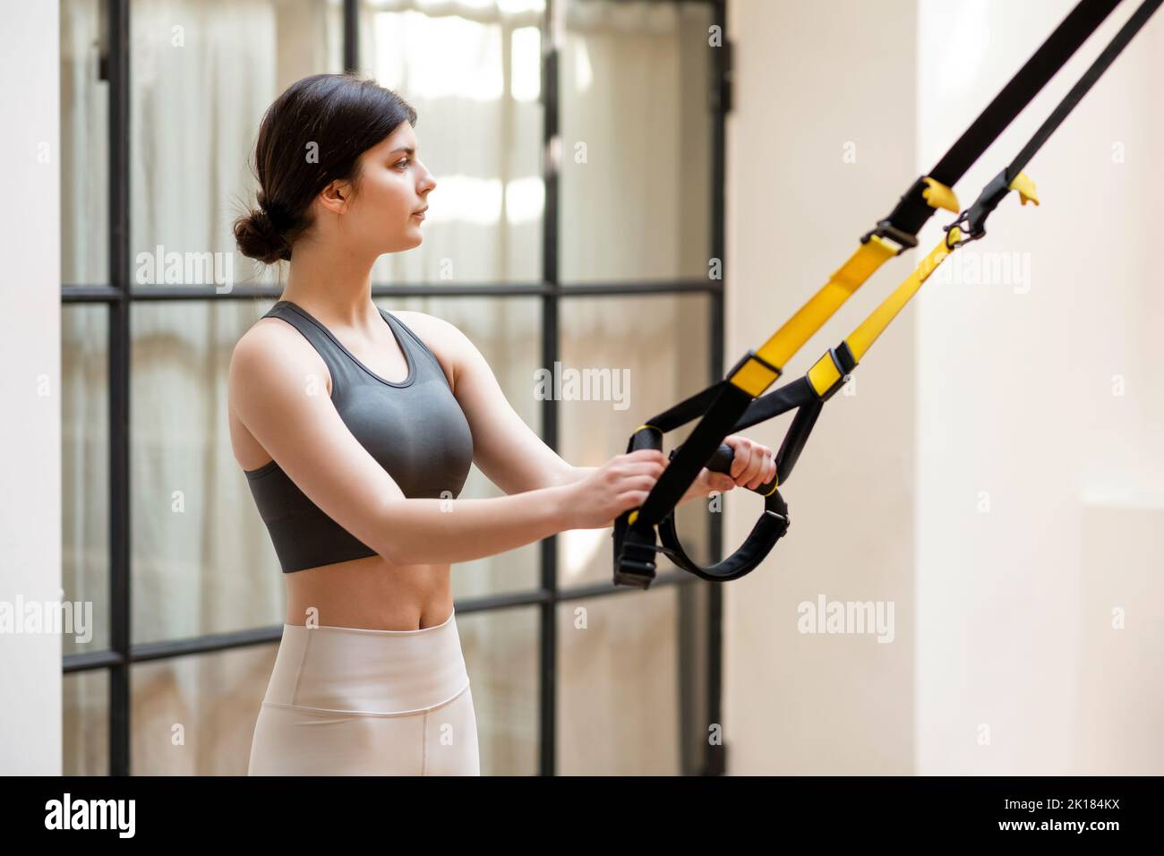 Seitenansicht einer jungen Sportlerin mit dunklem Haar im Crop-Top und Leggings, die an TRX-Loops ziehen und beim Training in einem modernen, hellen Fitnessstudio wegschauen Stockfoto