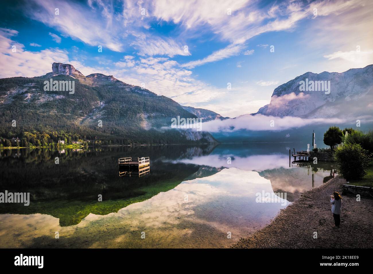 Atemberaubende Morgenansicht einer Frau, die am Ufer des Altausseer Sees, der Steiermark, Österreich, beim Kaffeetrinken und beim Blick auf den See steht Stockfoto