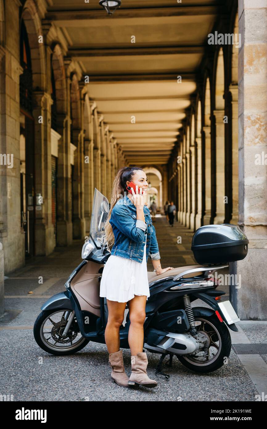 Junge lächelnde Frau auf einem Motorrad, die auf einem roten Smartphone spricht Stockfoto