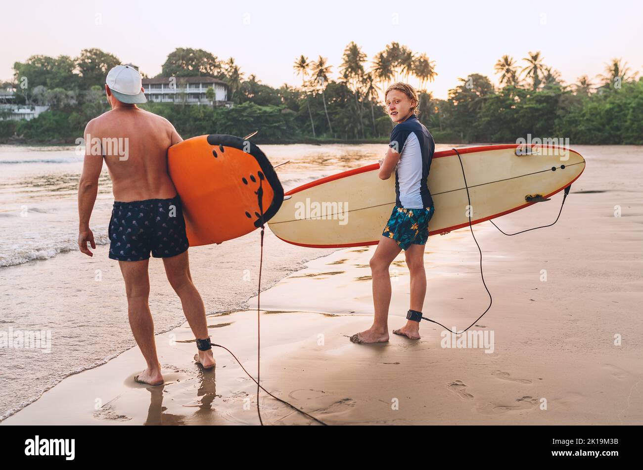 Teenager Junge mit Vater mit Surfbrettern gehen zum Surfen auf Sri Lanka Insel ins Meer. Sie haben einen Winterurlaub und genießen ein wunderschönes Sonnenuntergangslicht Stockfoto