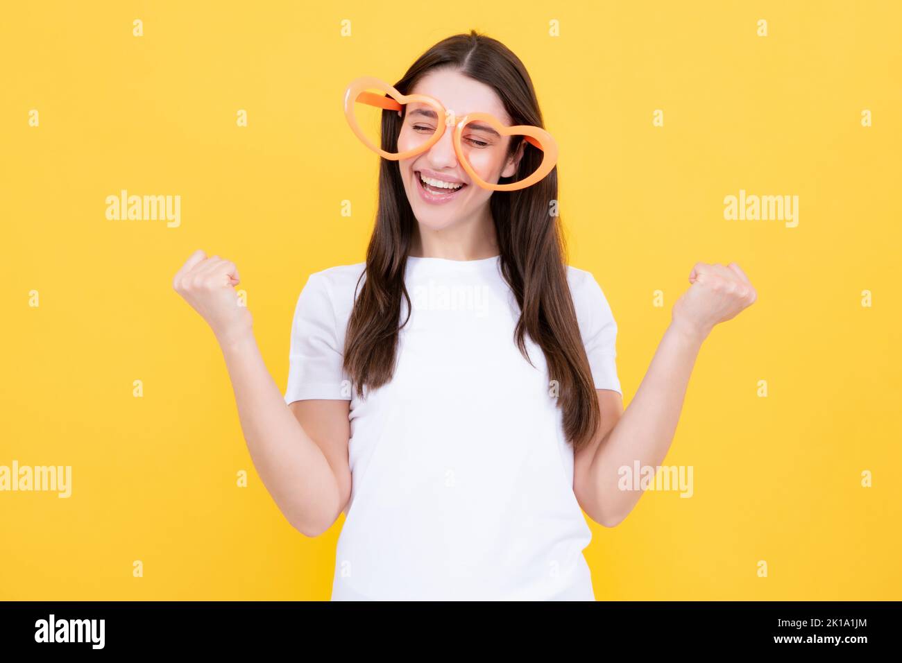 Frau fühlt sich cool und genial. Aufgeregt glücklich lustige junge Frau in Party-Brille in Form eines Herzens auf einem gelben hellen Hintergrund. Stockfoto