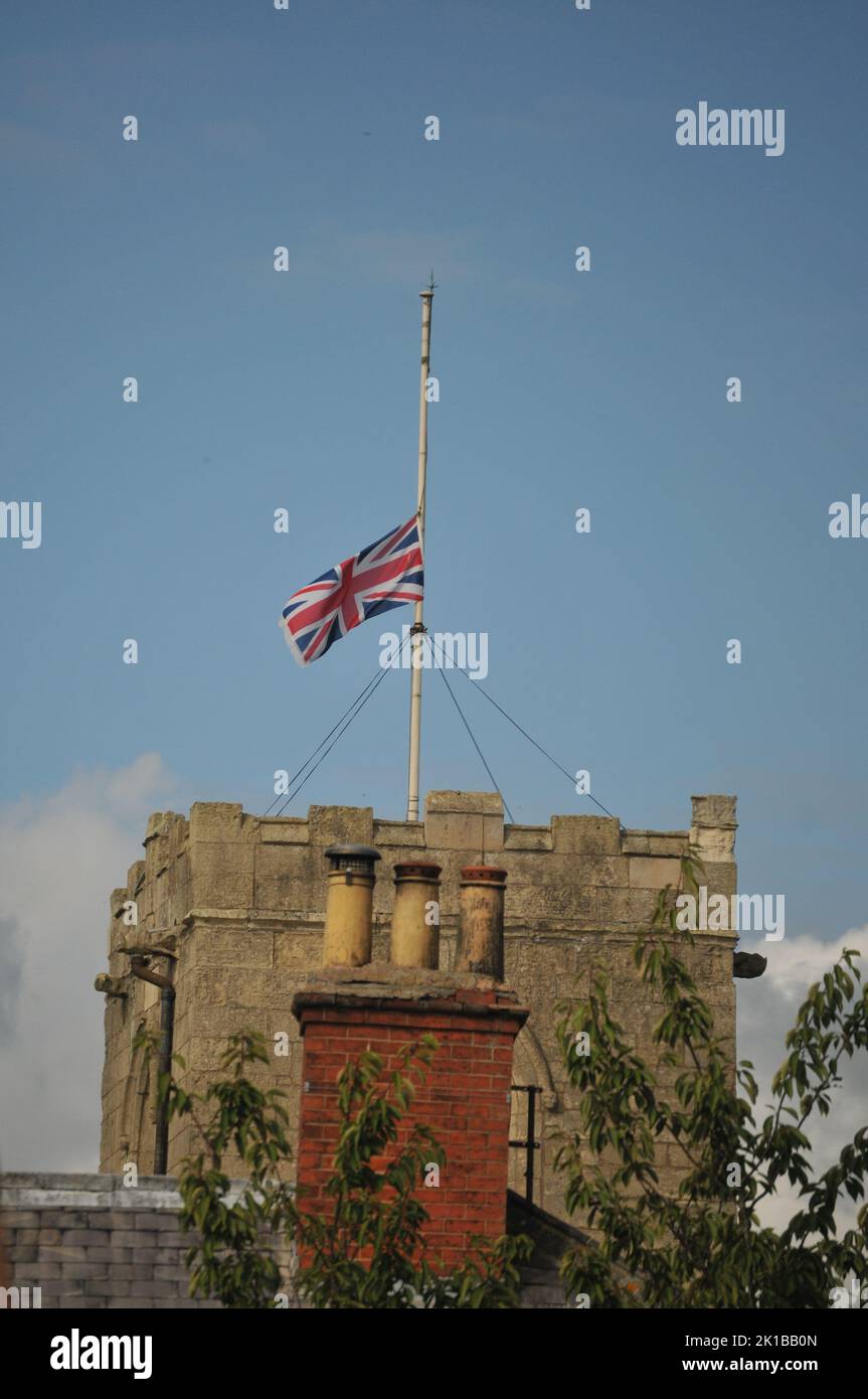 Kirche mit Flagge halb Mast Queen Elizabeth Tod. Blauer, sonniger Sommerhimmel und Nahaufnahme des Kirchturms und der Schornsteintöpfe. Stockfoto