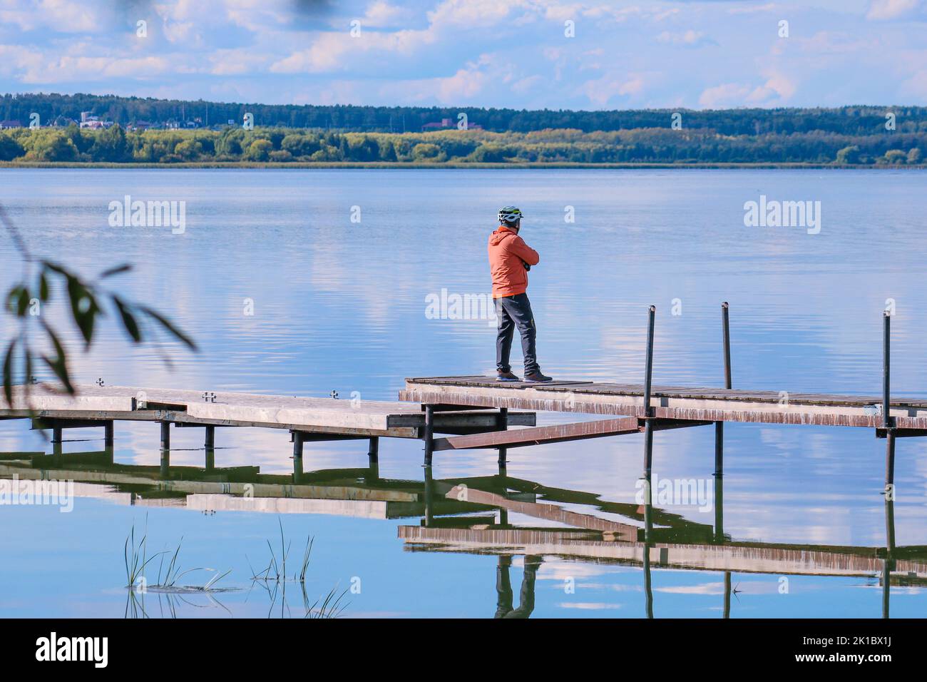 Ein unkenntlich männlicher Radfahrer in einem Helm steht auf dem Pier und bewundert die Oberfläche des Sees. Sport und Inspiration. Stockfoto