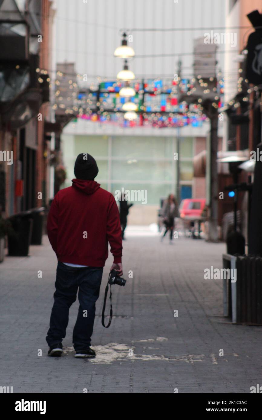 Eine vertikale Aufnahme eines Mannes mit einer Kamera in einem roten Hoodie, der mitten auf der Straße steht Stockfoto