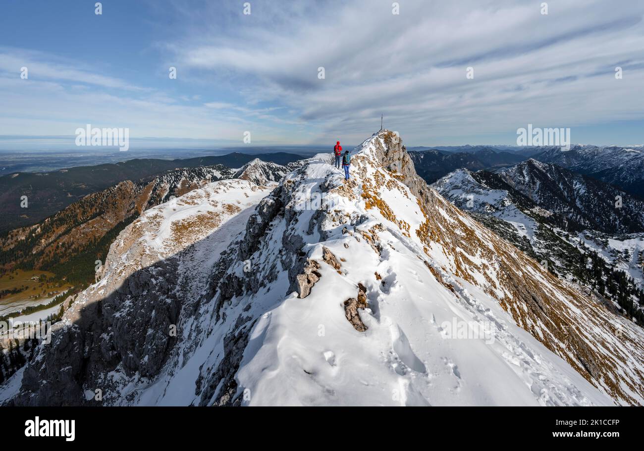 Zwei Bergsteiger auf einem schmalen, felsigen, schneebedeckten Bergrücken, im hinteren Gipfel der Ammergauer Hochplatte mit Gipfelkreuz, Blick auf das Bergpanorama, Wandern Stockfoto