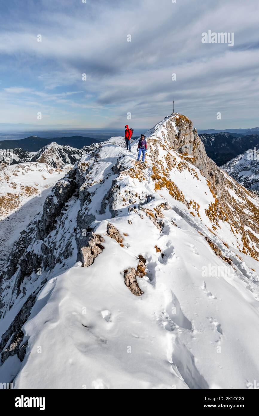 Zwei Bergsteiger auf einem schmalen, felsigen, schneebedeckten Bergrücken, im hinteren Gipfel der Ammergauer Hochplatte mit Gipfelkreuz, Blick auf das Bergpanorama, Wandern Stockfoto