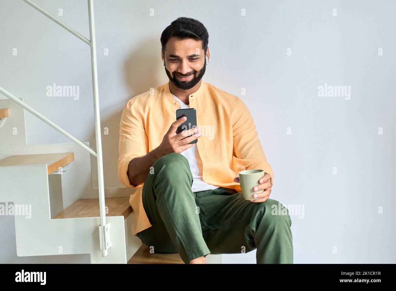 Glücklicher indischer Mann, der Ohrstöpsel trägt und Videos auf dem Smartphone zu Hause ansieht. Stockfoto