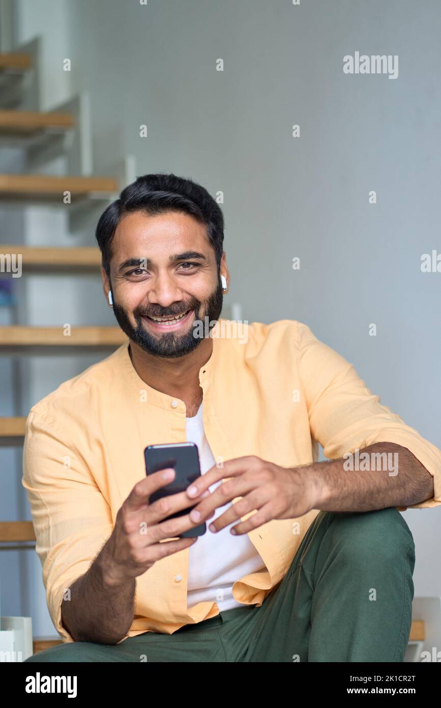 Glücklicher indischer Mann, der zu Hause in Ohrhörern sitzt und das Telefon senkrecht hält. Stockfoto