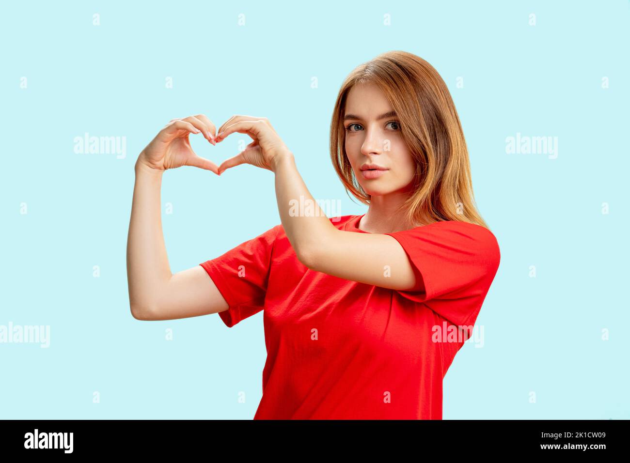 Unterstützendes Frauenporträt. Liebeszeichen. Dankbare zärtliche Dame im roten T-Shirt zeigt auf blauem Hintergrund eine vereinzelte Herzbewegung. Mitgefühl. Versicherungssanc Stockfoto