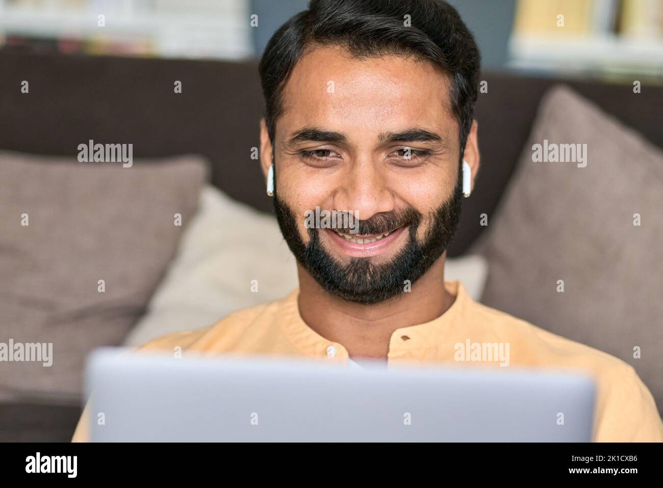 Lächelnder indischer Mann, der zu Hause mit Ohrhörern am Laptop sitzt. Stockfoto