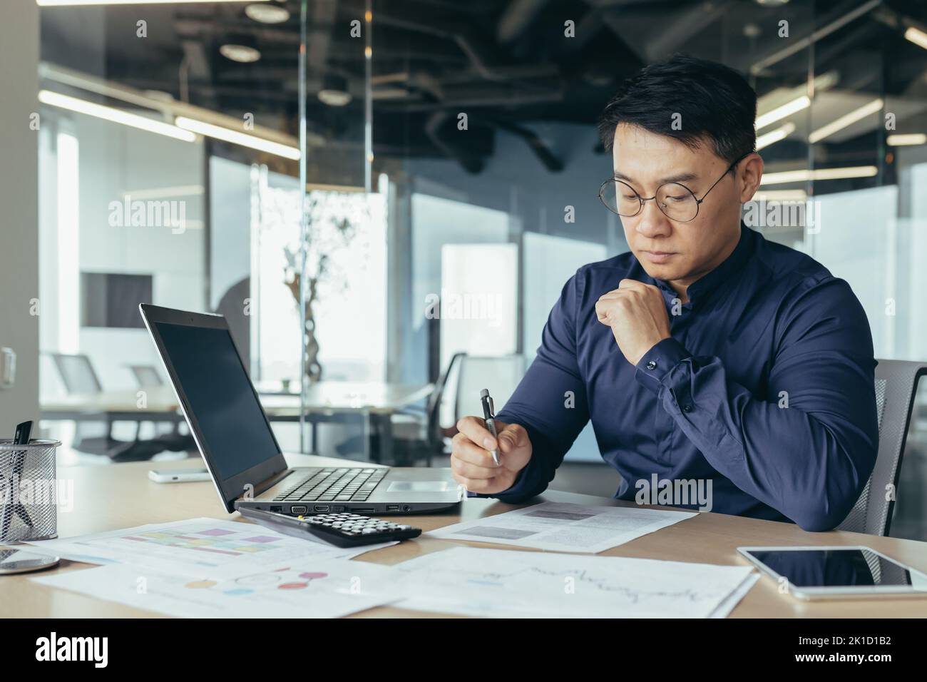 Denkender asiatischer Finanzier in Gläsern Buchhalter, der mit Dokumenten und Konten arbeitet, Laptop benutzt, männlicher Geschäftsmann, der Schreibarbeit beim Sitzen am Schreibtisch im Bürogebäude macht. Stockfoto