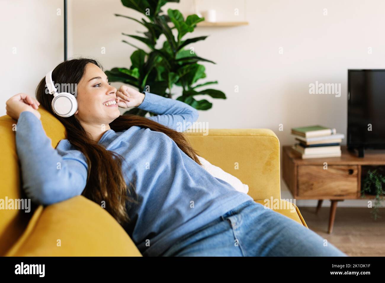 Entspannt lächelnde junge Erwachsene Frau mit Kopfhörern, die zu Hause auf dem Sofa sitzen Stockfoto