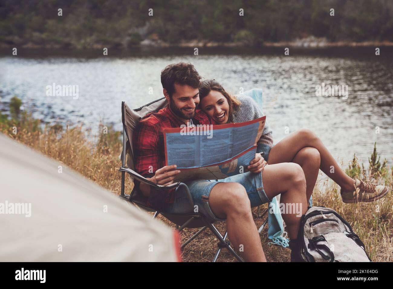 Abenteuerlustiges junges Paar, das sich einen Stadtplan anschaut, während es draußen auf Campingstühlen sitzt. Romantisches junges Paar Camping am See. Stockfoto